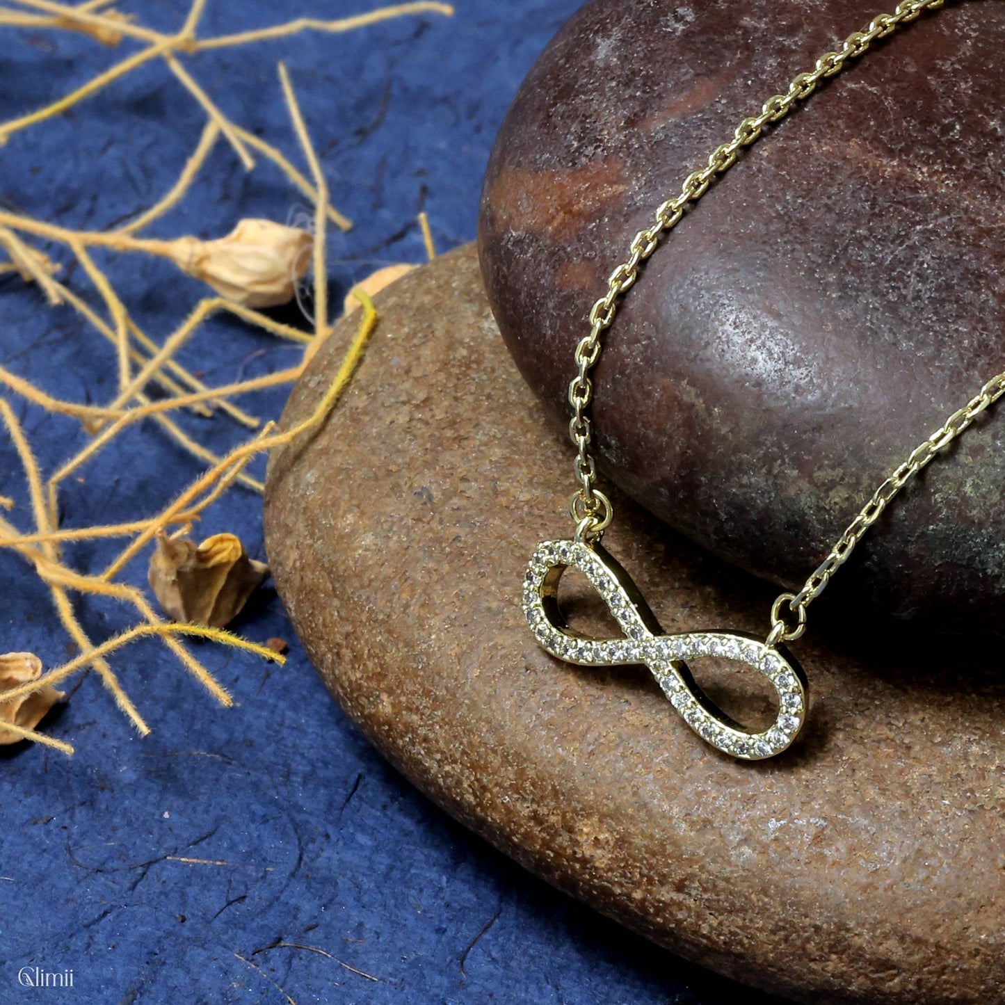 Glimii dainty infinity pendant necklace with sparkling stones on a gold chain displayed on smooth stones