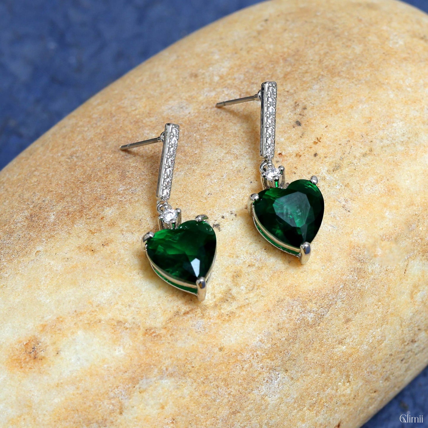 Glimii green heart drop earrings with silver settings and sparkling stones on a beige stone background