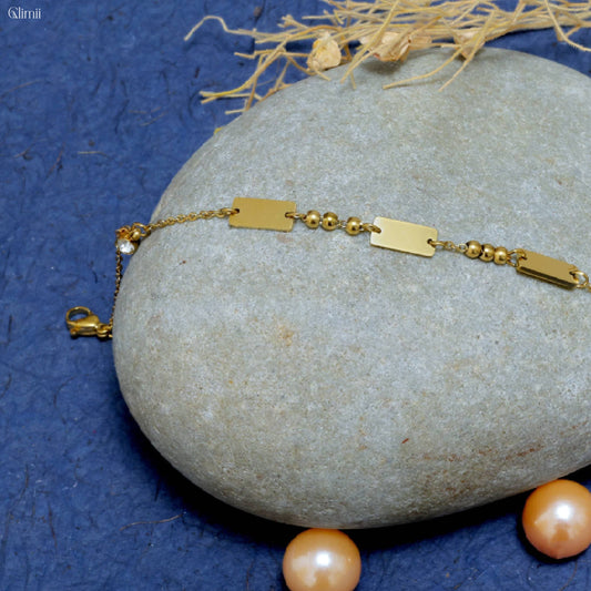 Glimii gold minimalist bracelet with rectangular links and small beads displayed on a smooth gray stone