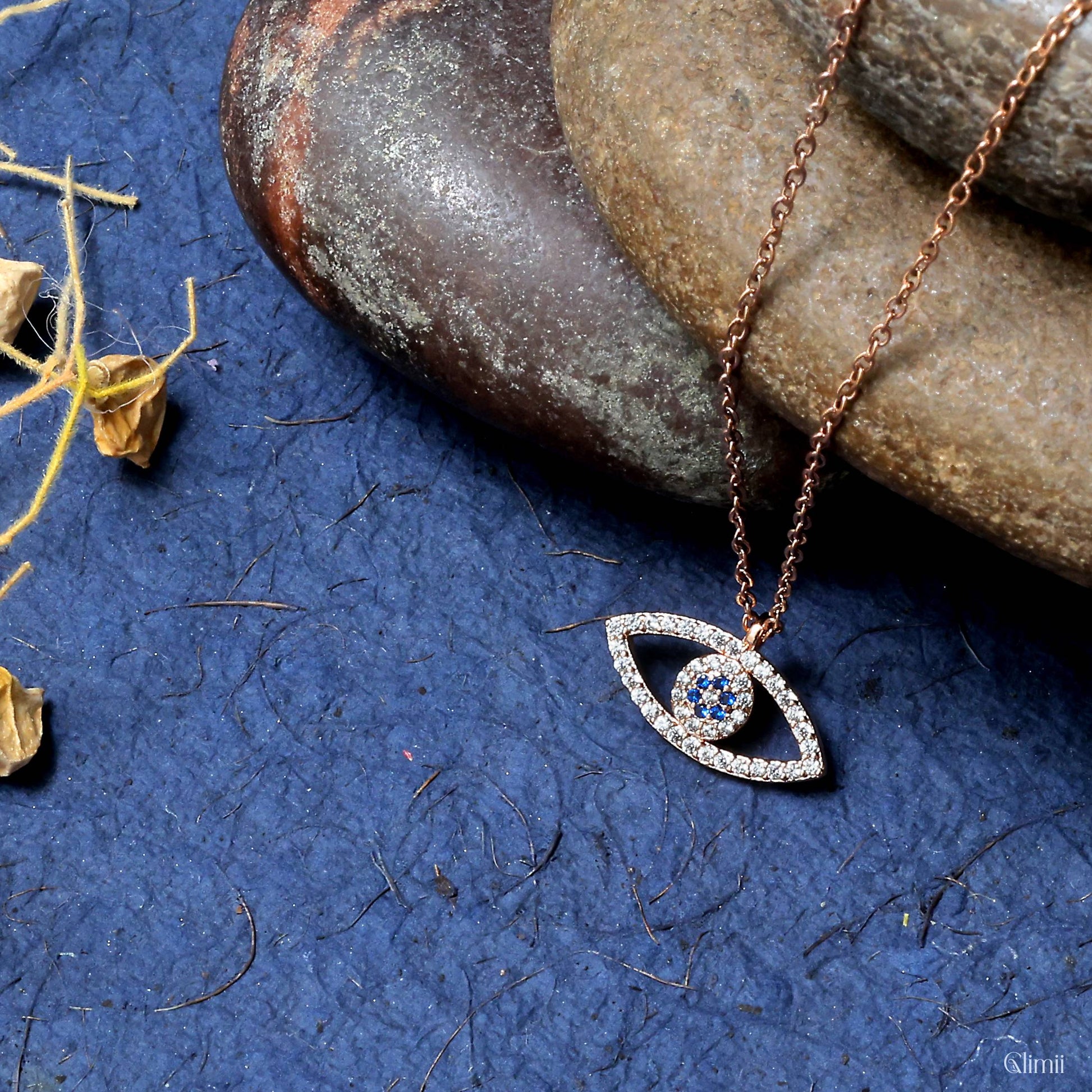 Glimii evil eye necklace with blue and clear stones on rose gold chain displayed on textured blue surface with stones