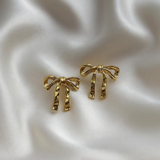 Glimii gold bow stud earrings with hammered texture displayed on white satin fabric