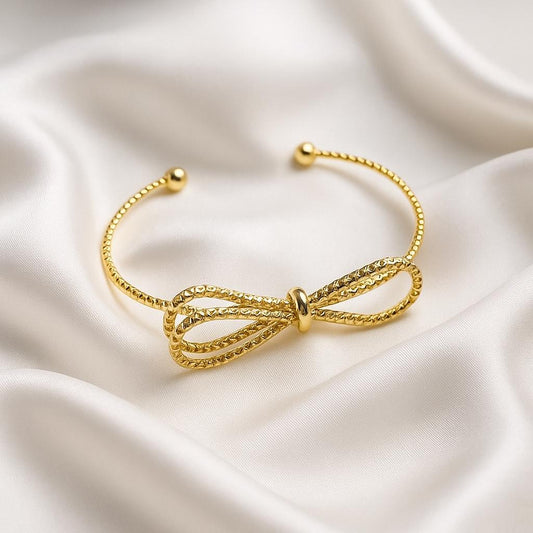 Glimii gold plated bow bracelet with textured design on a satin fabric background