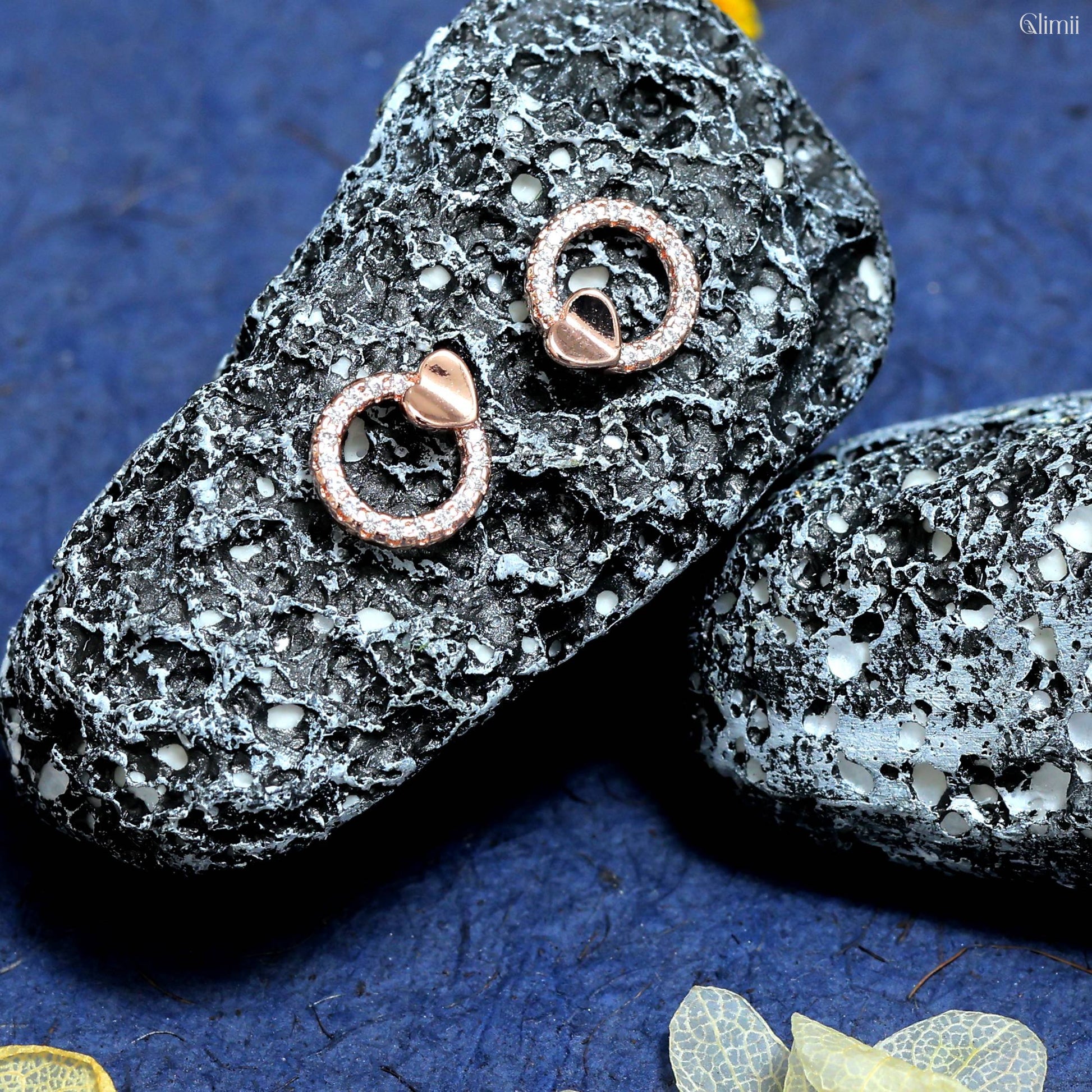 Glimii minimalist heart earrings in rose gold on textured black stones with blue background